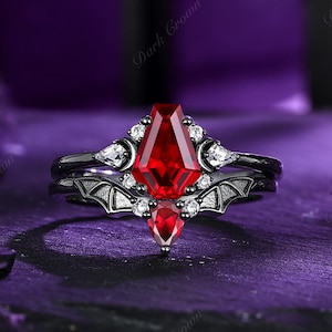 May include: A black ring set with a large, faceted, red gemstone. The ring features a bat wing design and is accented with smaller white gemstones. The words "Dark Crown" are visible in the image.