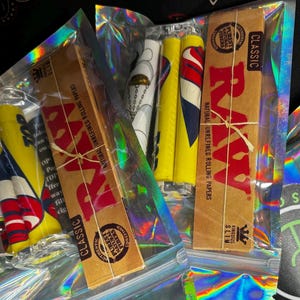 Könnte beinhalten: Verschiedene Raucheraccessoires, darunter RAW Classic Zigarettenpapier, vorgedrehte Hülsen und Feuerzeuge, verpackt in transparenten, irisierenden Beuteln. Die RAW-Papiere sind mit Bindfaden verschnürt.
