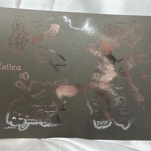 Puede incluir: Un mapa de la tierra ficticia de Erilea, impreso en un fondo gris oscuro con acentos plateados y oro rosa. El mapa presenta varias regiones y ubicaciones, incluyendo el "Mar de Ono" y "Las Tierras Soñadas".
