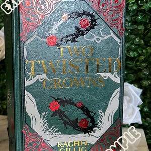 May include: A green hardcover book with the title "Two Twisted Crowns" in gold lettering. The book cover features a black and white illustration of a thorny crown with red roses. The author's name, "Rachel Gillig", is printed in gold lettering at the bottom of the cover.
