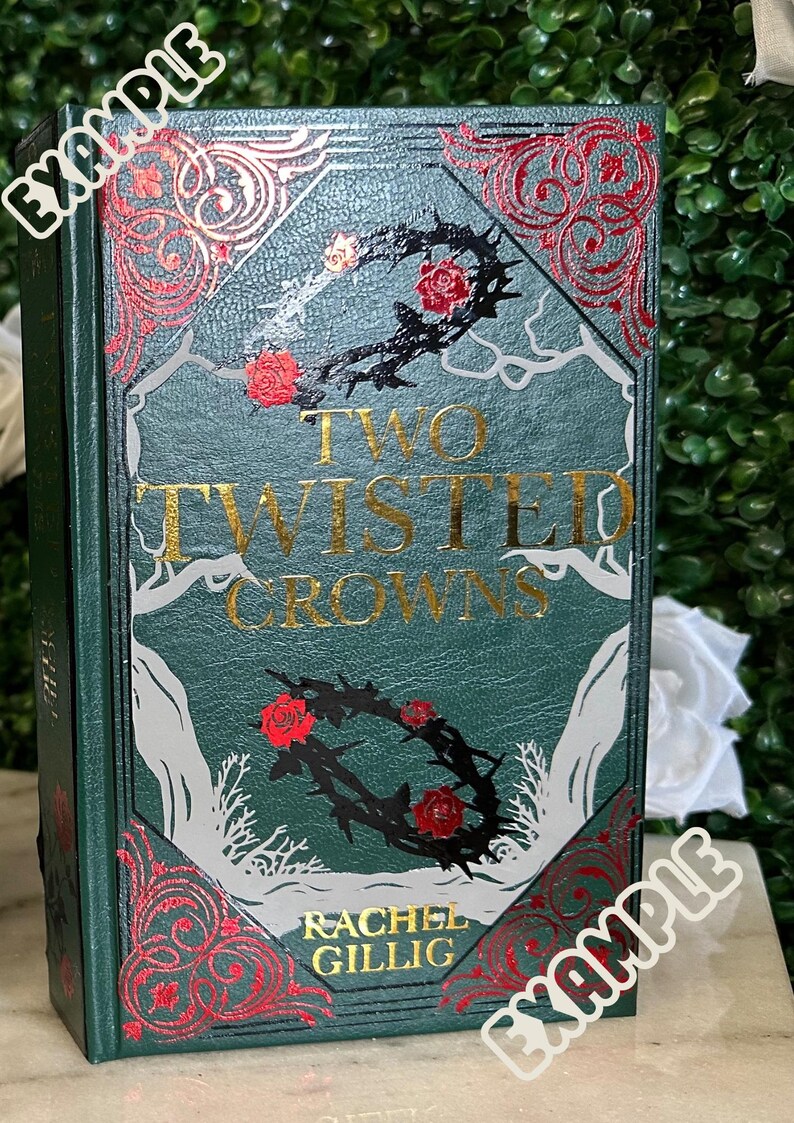 May include: A green hardcover book with the title "Two Twisted Crowns" in gold lettering. The book cover features a black and white illustration of a thorny crown with red roses. The author's name, "Rachel Gillig", is printed in gold lettering at the bottom of the cover.