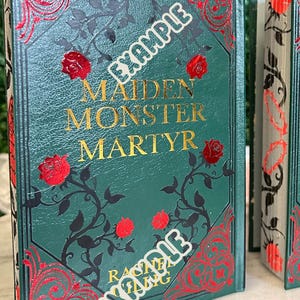 May include: A green hardcover book with gold lettering that reads "Maiden Monster Martyr" by Rachel Gillig. The book cover features a black and red floral design with roses.