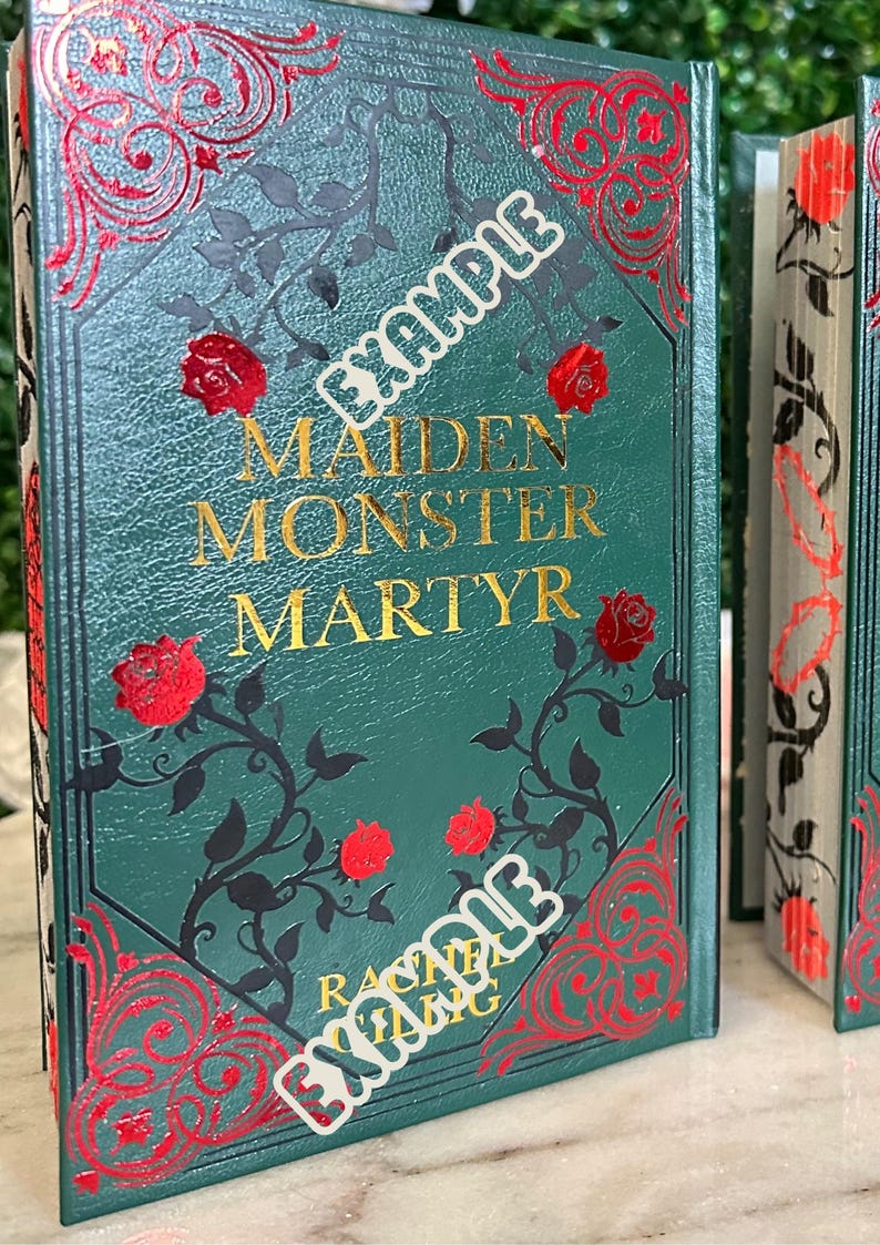 May include: A green hardcover book with gold lettering that reads "Maiden Monster Martyr" by Rachel Gillig. The book cover features a black and red floral design with roses.