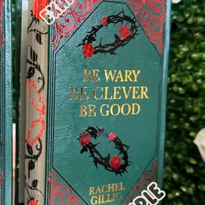May include: A green hardcover book with gold lettering that reads "Be wary, be clever, be good." The author's name, Rachel Gillig, is printed below the text. The book cover features a black and red floral design.
