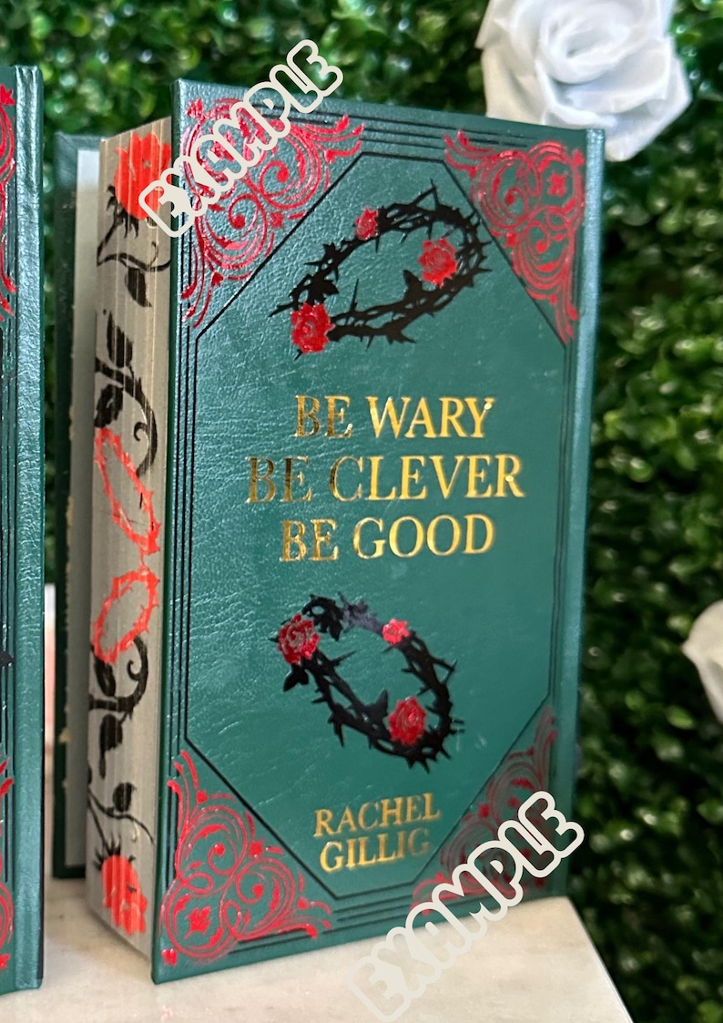 May include: A green hardcover book with gold lettering that reads "Be wary, be clever, be good." The author's name, Rachel Gillig, is printed below the text. The book cover features a black and red floral design.