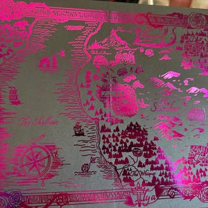 May include: A pink foil map with a detailed design of a fictional land. The map includes names like "The Shallow", "The Whisper", and "Ilya".