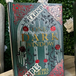 May include: A green hardcover book with gold lettering that reads "One Dark Window" by Rachel Gillig. The book cover features a decorative design with red and gold accents.