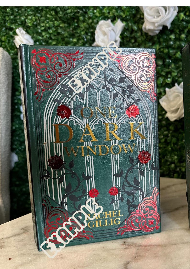 May include: A green hardcover book with gold lettering that reads "One Dark Window" by Rachel Gillig. The book cover features a decorative design with red and gold accents.