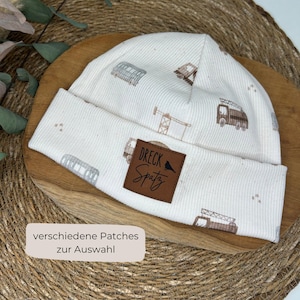 Könnte beinhalten: Cremefarbene Mütze mit einem sich wiederholenden Muster aus Baufahrzeugen und Gebäuden. Ein braunes Lederpatch mit dem Text "Dreck Spatz" ist auf der Vorderseite aufgenäht. Der Text "verschiedene Patches zur Auswahl" befindet sich in einem weißen Rechteck.