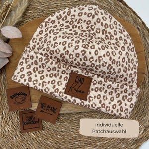 Könnte beinhalten: Eine beige-braune Beanie-Mütze mit Leopardenmuster und umgeschlagenem Saum. Die Mütze hat einen braunen Lederaufnäher mit dem Text "CIAO Kakao". Zusätzliche Lederaufnäher mit Text und Grafiken sind ebenfalls vorhanden.