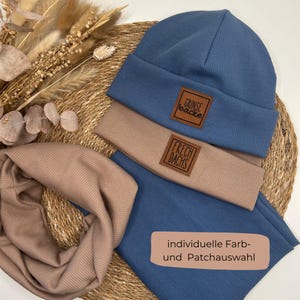 Könnte beinhalten: Ein blaues geripptes Strickmützen- und Halstuch-Set mit braunen Lederaufnähern. Die Mütze trägt den Text "Grinse Backe" und das Halstuch den Text "Frech Dachs". Das Set wird auf einer gewebten Matte präsentiert.