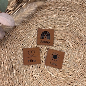 Könnte beinhalten: Drei braune Lederquadrate mit schwarzen Grafiken und Text. Ein Quadrat hat einen Regenbogen und den Namen "Emilio". Ein anderes hat ein Herz und den Namen "Mina". Das dritte hat eine Sonne und den Namen "Leon".