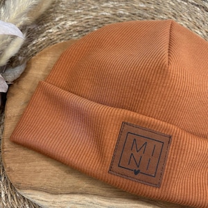Könnte beinhalten: Ein rostfarbener Rippstrick-Beanie mit einem quadratischen Lederaufnäher. Der Aufnäher zeigt die Buchstaben "MI NI" in einem minimalistischen Design mit einem Herzsymbol. Der Hut ist aus weichem, warmem Material.