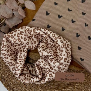 Könnte beinhalten: Ein Haargummi mit Leopardenmuster in Braun und Creme. Daneben ein beiger Stoff mit schwarzen Herzformen. Das Haargummi liegt auf einer gewebten Matte. Das Bild enthält auch den Text "große Stoffauswahl".