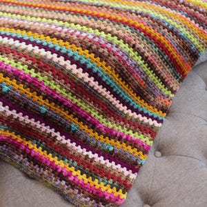May include: A colorful striped crocheted throw blanket with a variety of colors, including pink, purple, green, yellow, and brown. The blanket is folded and draped over a gray tufted ottoman.