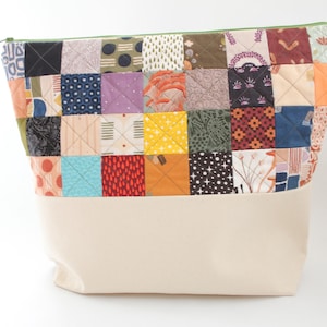 May include: A patchwork fabric zippered pouch with a cream-colored base. The upper section features a variety of colorful, patterned squares in shades of blue, orange, yellow, and green. The zipper is green.