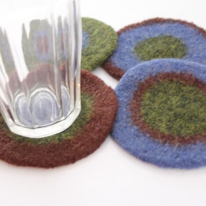 May include: Four round knitted coasters with a brown, green, and blue color scheme. The coasters are made of wool and have a fuzzy texture.