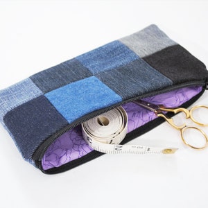 May include: A denim patchwork pencil case with a black zipper, open to reveal a measuring tape and gold scissors. The interior lining is purple with a floral pattern. The case is rectangular.