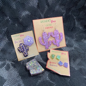 May include: Three sets of purple and green glitter cactus earrings and pins. The earrings and pins are labeled "Desert Flair Cactus Earrings" and "Desert Flair Cactus Pin Set".
