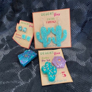 May include: A set of three turquoise glitter cactus earrings, a turquoise glitter cactus pin, and a purple glitter flower pin. The earrings and pins are all handmade and have a desert flair theme. The cards are labeled "Desert Flair Cactus Earrings" and "Desert Flair Cactus Pin Set".