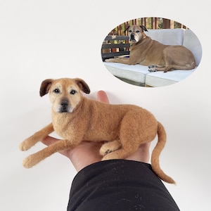 Op de afbeelding: Een handgemaakte, bruin gekleurde hondensculptuur met donkerbruine oren en een gedetailleerd gezicht. De hond ligt uitgestrekt, met zijn poten uitgestrekt. Een ovale afbeelding toont een echte hond op een witte bank.