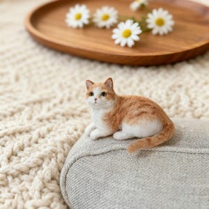 May include: A handmade orange and white cat figurine resting on a gray cushion. The cat is made of wool and has detailed features. Daisies and a wooden tray are in the background, creating a cozy, natural aesthetic.