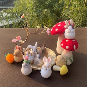 May include: A collection of handmade felted bunnies in various colors, including white, brown, and gray, arranged with a red mushroom with white spots and a small orange carrot. A decorative branch with pink flowers is also included.