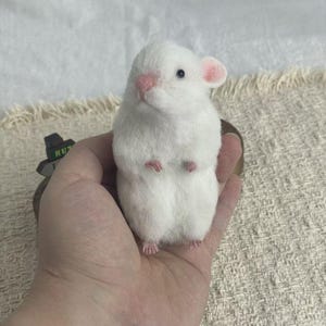 以下が含まれることがあります： 柔らかくふわふわした質感の、白いニードルフェルトで作られた小さなハムスターのフィギュア。ハムスターは淡いピンクの鼻、小さなピンクの耳、そして小さなピンクの足を持っています。暗い丸い目を持ち、直立した姿勢で立っています。