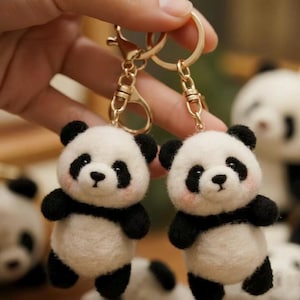 Könnte beinhalten: Zwei Panda-Bär-Schlüsselanhänger. Jeder Panda hat einen weißen Körper, schwarze Arme, Beine und Ohren sowie schwarze Markierungen um die Augen. Die Pandas haben rosige Wangen und sind an goldfarbenen Schlüsselringen befestigt.