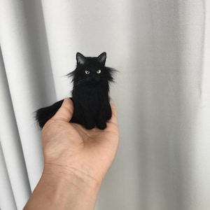 May include: A miniature black cat figurine with long, fluffy fur, pointed ears, and bright yellow-green eyes. The small, detailed sculpture features a realistic feline appearance with a long tail and is crafted with a soft, textured finish.