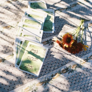 May include: A stack of green and white wedding invitations with calligraphy that reads "Blair & Afton married at home, in the garden Saturday April 13, 2019". The invitations are on a white tablecloth with a floral pattern.