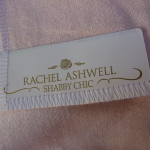 May include: A close-up of a white label with the text "RACHEL ASHWELL SHABBY CHIC" in gold lettering. The label is stitched onto a light pink fabric. A rose graphic is above the text.