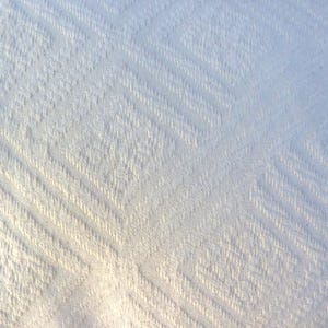 May include: Close-up of a white fabric with a textured, diagonal pattern. The fabric has a subtle, light-reflecting quality, with variations in tone creating depth and visual interest. The pattern is consistent across the surface.