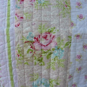 May include: A close-up of a quilted fabric with a floral pattern. The quilt features a patchwork design with vertical stripes of varying widths. The color palette includes pink, white, green, and light blue, with a focus on rose and floral motifs.