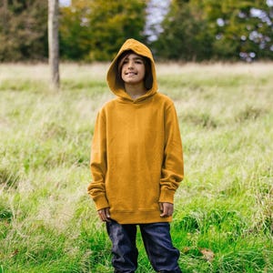 Pode incluir: Uma pessoa jovem vestindo um moletom amarelo mostarda com capuz de cordão. A pessoa está em pé em um campo de grama alta.