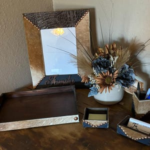 May include: A set of five western-themed home decor items, including a tray, a mirror, a small box, a pen holder, and a card holder. The items are made of wood and have a brown and white cowhide pattern. The mirror has a brown frame with a gold-colored inscription that reads "Family".