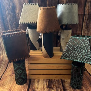 May include: Five table lamps with different colored and patterned lampshades. The lampshades are made of leather and have a stitched edge. The bases of the lamps are also made of leather and have different patterns. The lamps are arranged on a wooden surface.
