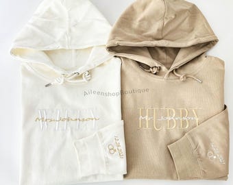 May include: Two hooded sweatshirts, one white and one beige, with embroidered text. The white sweatshirt says "WIFEY" with a gold ring symbol and the date "12.28.2021". The beige sweatshirt says "HUBBY" with a gold ring symbol and the date "12.28.2021".