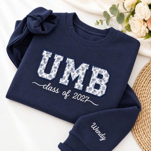 Pode incluir: Uma camisola azul-marinho de gola redonda com as letras UMB num padrão floral branco e azul. Abaixo das letras, o texto diz class of 2027 em letra branca. A manga está bordada com o nome Wendy em letra cursiva branca.