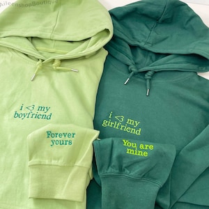 May include: Two green hoodies with drawstrings. One is light green with "i <3 my boyfriend" embroidered on the front and "Forever yours" on the sleeve. The other is dark green with "i <3 my girlfriend" and "You are mine" embroidered.