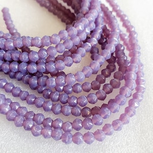 Puede incluir: Una hebra de cuentas de vidrio de color lavanda con un diseño facetado en forma de estrella. Las cuentas están enhebradas en un hilo transparente.