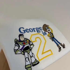 Może przedstawiać: Biała tkanina z haftowanymi postaciami Buzza Astrala i Chudego z Toy Story, wraz z napisem "George is 2" w kolorze niebieskim i ż&oacute;łtym. Buzz Astral jest w klasycznej pozie, a Chudy macha.