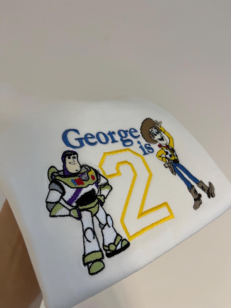 Może przedstawiać: Biała tkanina z haftowanymi postaciami Buzza Astrala i Chudego z Toy Story, wraz z napisem "George is 2" w kolorze niebieskim i ż&oacute;łtym. Buzz Astral jest w klasycznej pozie, a Chudy macha.