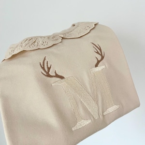 Puede incluir: Babero beige para bebé con cuello de encaje decorativo. El babero presenta un diseño de cuernos bordados en marrón y la letra "M" en hilo beige a juego. El babero está hecho de tela suave.