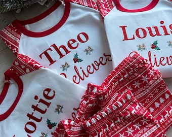 May include: Three white Christmas pajamas with red trim and red and white patterned pants. The pajamas are personalized with the names "Theo", "Lottie", and "Louis" and the phrase "Believe".