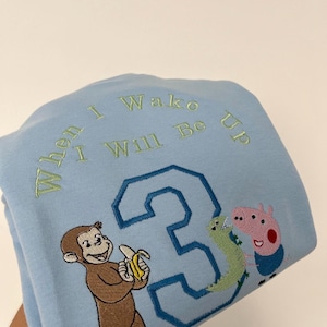 May include: Pale blue sweatshirt with embroidered text "When I Wake I Will Be Up" and the number "3". Features a cartoon monkey holding a banana, a green dinosaur, and a pink pig.