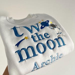 May include: White sweatshirt with blue embroidered text that reads "Two the Moon" and a small astronaut graphic. The name "Archie" is embroidered below the text.