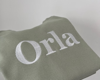 Könnte beinhalten: Gefalteter, salbeigrüner Waffelstrickstoff mit dem Namen "Orla" in Weiß gestickt. Der Text ist in einer klaren, serifenlosen Schriftart gehalten. Die Stoffstruktur ist deutlich sichtbar.