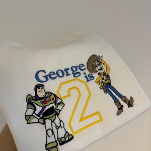 Może przedstawiać: Biała bluza z haftowanymi postaciami Buzza Astrala i Chudego z Toy Story. Tekst "George ma 2 lata" jest wyhaftowany w kolorze niebieskim i ż&oacute;łtym. Kolorowy design na urodziny dziecka.