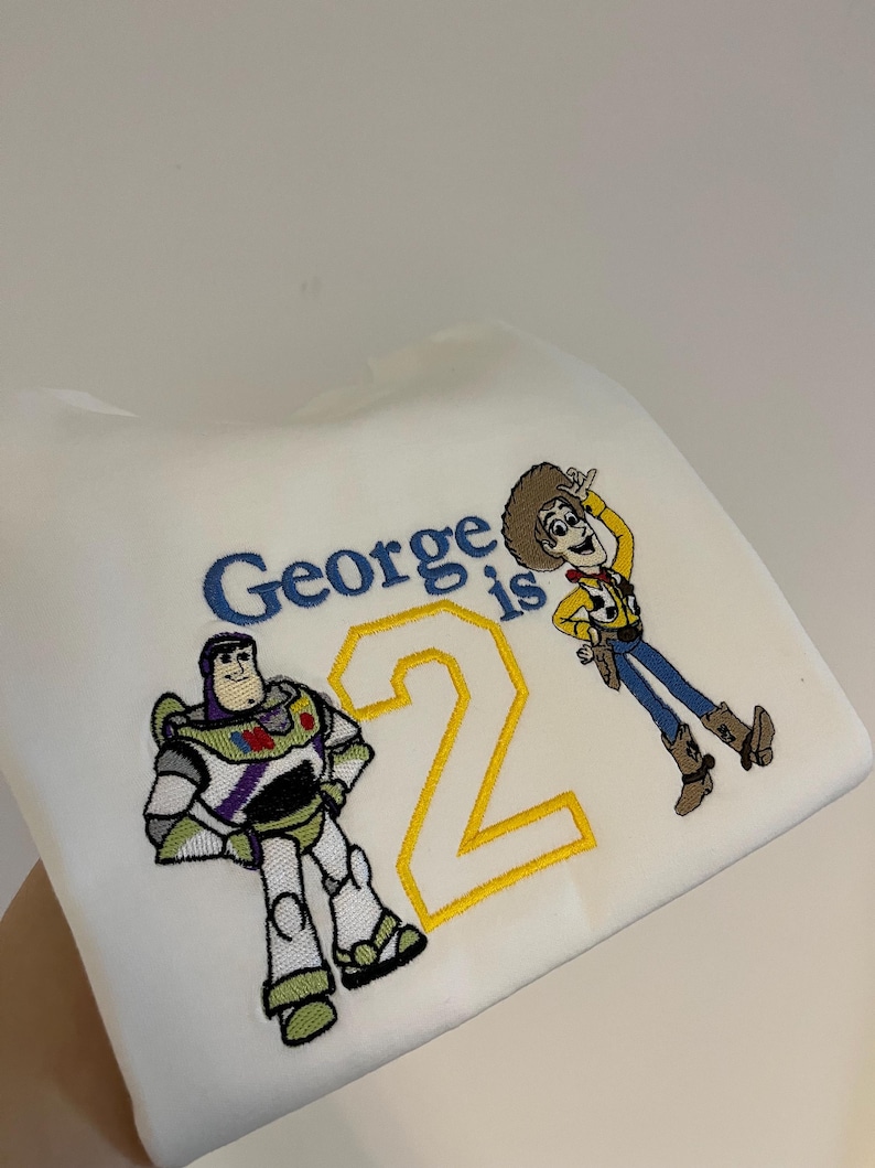 Może przedstawiać: Biała bluza z haftowanymi postaciami Buzza Astrala i Chudego z Toy Story. Tekst "George ma 2 lata" jest wyhaftowany w kolorze niebieskim i ż&oacute;łtym. Kolorowy design na urodziny dziecka.